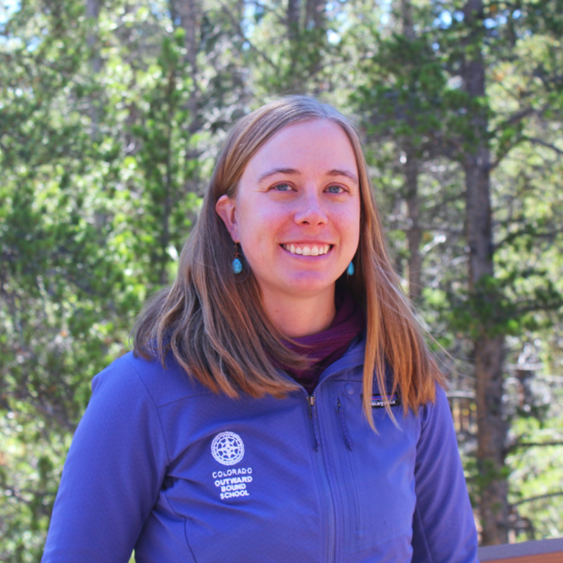 The image shows a woman with blonde hair, wearing a purple jacket with a "Colorado Outward Bound School" logo. She is smiling and has turquoise earrings. The background features a forest with green trees, suggesting an outdoor setting. The photo appears to be taken on a sunny day.